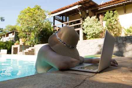 middle-aged woman working on the laptop inside the pool on very hot summer day, almost giving up workの写真素材