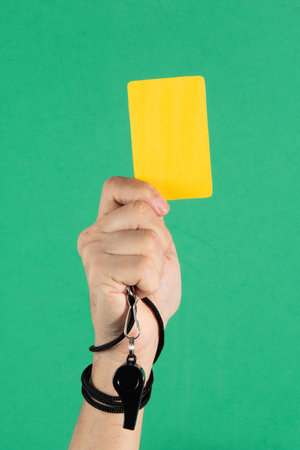 referees hand holding a yellow card and whistle on green background.