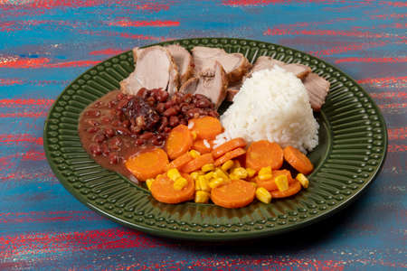 Typical Brazilian food dish with rice, red beans, pork and carrot salad and braised corn in a green plateの写真素材