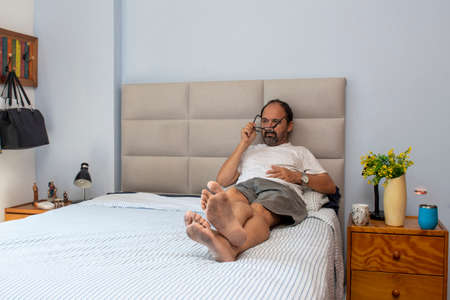 gentleman sitting on bed in bedroom relaxing and taking off his glassesの写真素材