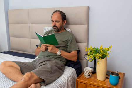 gentleman sitting on bed in bedroom reading literature bookの写真素材