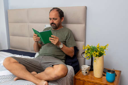 gentleman sitting on bed in bedroom reading literature bookの写真素材