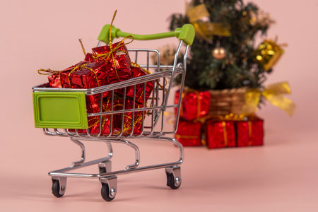 Christmas shopping cart with gifts and Christmas tree in background on light red background, selective focusの写真素材
