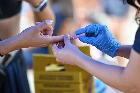 A nurse pricks the patient's finger to perform a blood glucose test. Disposal box in the background.