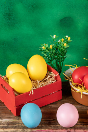 Colorful Easter eggs arranged in a red wooden crate and small bowl with straw, placed on a rustic...