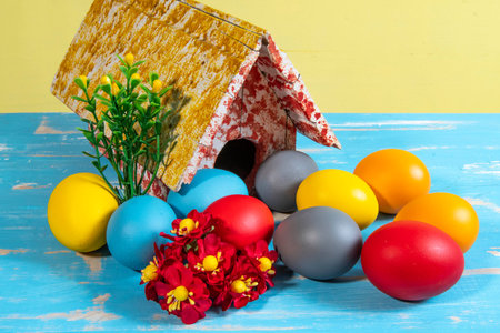 Colorful Easter eggs arranged with flowers and greenery beside a rustic decorative house on a blue...