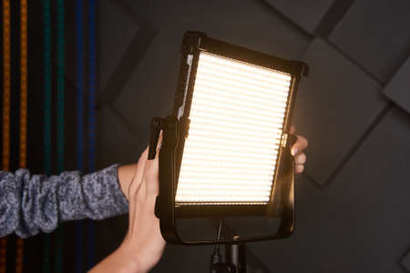 studio assistant fix professional video light on adjustable light stand. Led lamp on tripod in photo studio. Photo and video lamp on embossed grey wall background. Close-up viewの写真素材