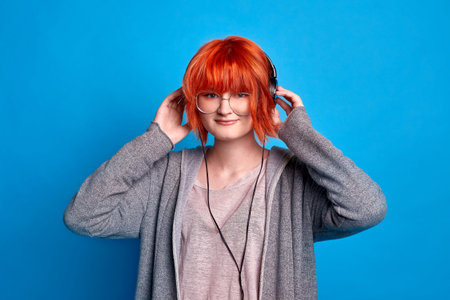 teenage girl in headphones listens to music on blue background. girl with bright red hair in anime...