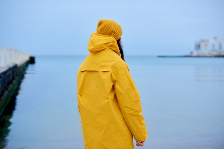 loneliness woman in yellow rain coat walking coast. Woman standing alone on beach and look at distant cityの写真素材
