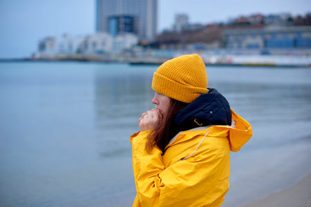 lonely woman in yellow raincoat freezes on sea shore against backdrop of winter cityscape in cold...