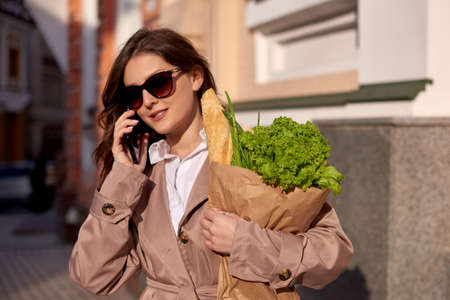 young woman talking on phone and holding a package of groceries in her hands walking down the streetの写真素材
