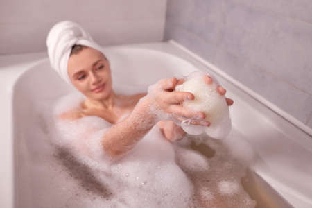 close up woman hands with shower sponge in foam bathtub at home. white towel in form of turban on head. shallow depth of field photoの写真素材