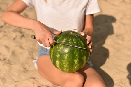 girl holds and cut with a knife whole ripe green watermelon in her hands. picnic on the beach. young woman in straw hat with long legs sit on sand in summer vacationの写真素材