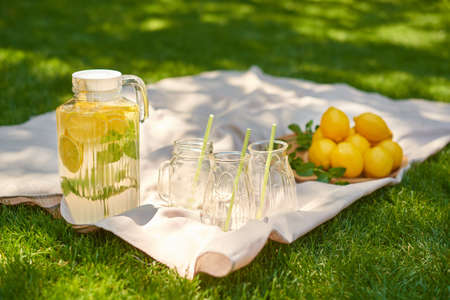 summer lemonade picnic with glass jug of cool drink on light bedding on green grassの写真素材