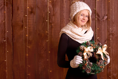 girl with christmas tree wreath decorate wooden wall at home. young woman in winter knitted clothes on brown wooden backgroundの写真素材
