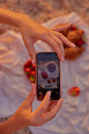 woman hands taking photo flatlay of summer picnic with croissants and wine glasses on beach with smartphoneの写真素材