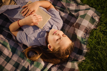 child girl hugs book lying down on checkered plaid on green grass in summer picnic in parkの写真素材
