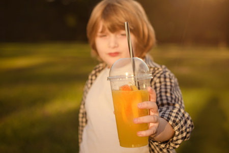 unhappy young boy holding plastic glass with orange juice or smoothie in park. focus on glass. face out of focusの写真素材