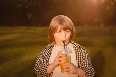 serious child boy drink juice through tube and holding plastic glass with orange smoothie in parkの写真素材