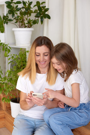 young mother and daughter use smartphone and smile while sitting on sofa at home. family time...