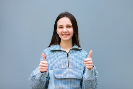 teen girl doing happy thumbs up gesture with both hands and looking at camera on blue backgroundの写真素材