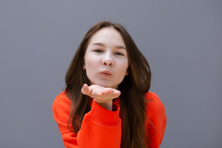 teen girl in red sport sweatshirt blows an air kiss and looking at camera on gray wall backgroundの写真素材