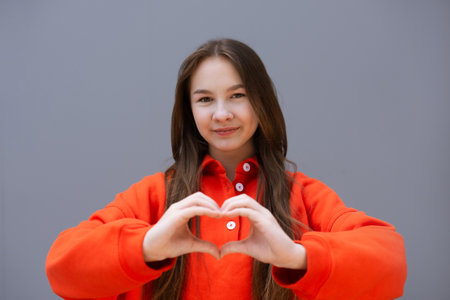 teen girl in red sport sweatshirt show heart shape gesture and smile on gray wall backgroundの写真素材