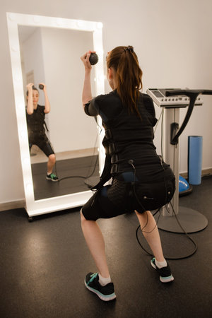 Sporty woman in electrical muscular stimulation suit doing exercise in gym
