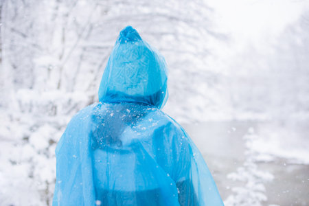 woman in blue raincoat walking through snowy forest near frozen lakeの写真素材