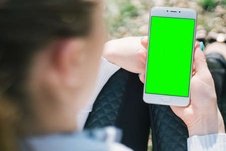 Girl with smartphone in hands with mock up green screen of blank screen sits in park on open space, screen for content integration. Hands holding gadget on blurred backdrop, front viewの写真素材
