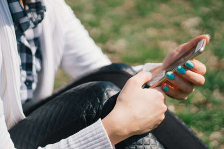 Beautiful young European girl sits on the grass in the park and uses a smartphone, concepts of using gadgets in a natural environment in the fresh airの写真素材