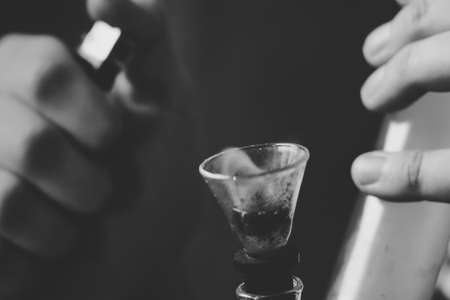 bong and a lighter in his hands. A man smokes cannabis weed, Smoke on a black background. Concepts of medical marijuana use and legalization of the cannabis. On a black backgroundの写真素材