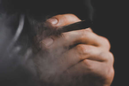 A man smokes weed cannabis a joint and a lighter in his hands. Smoke on a black background. Concepts of medical marijuana use and legalization of the cannabis. On a black backgroundの写真素材