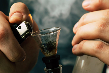 bong and a lighter in his hands. A man smokes cannabis weed, a Smoke on a black background. Concepts...