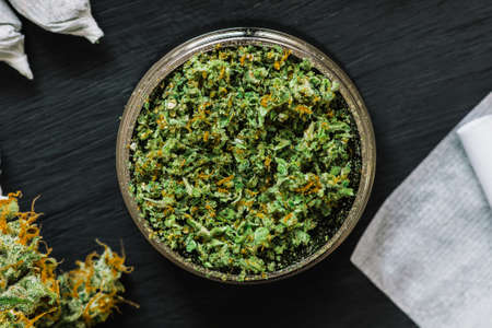 Grinder for chopping cannabis flowers with fresh grass surrounded by jambs and paper for a self-rolled on a black background woodenの写真素材