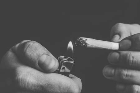 A man smokes cannabis weed, a joint and a lighter in his hands. Smoke on a black background. Concepts of medical marijuana use and legalization of the cannabis. On a black backgroundの写真素材