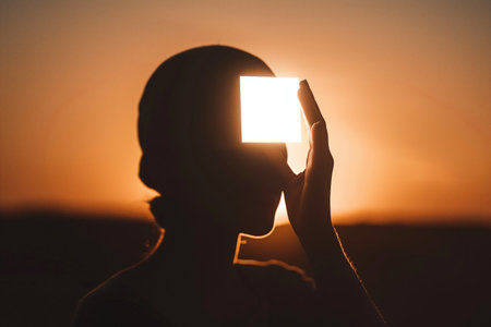 The silhouette of an individual holding a glowing box in front of their face against a vivid sunsetの素材