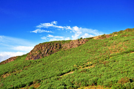 Red rock  surrounded by green vegetation under blue cloudy sky at Holyrood park in Edinburgh, Scotlandの写真素材