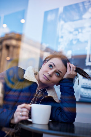 Young pretty girl sitting inside a modern cafe drinking coffee or tea. Cute Caucasian model. View...