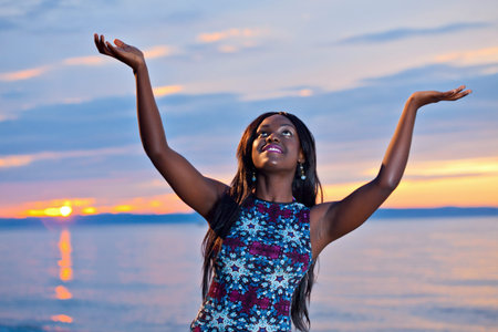 Portrait of beautiful black African American woman posing with open hands up to the sky on the beach at sunsetの写真素材