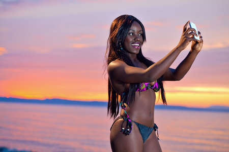 Portrait of beautiful black African American woman posing to take a selfie with her smartphone on the beach at sunsetの写真素材