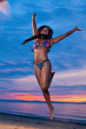 Beautiful black African American woman jumping up the air on the beach at sunsetの写真素材