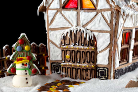 Sugar snowman stands next to decorative gingerbread house with lights inside on black background. Rural Christmas night sceneの写真素材