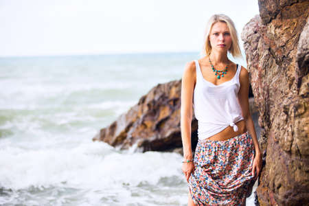 Beautiful young blonde woman posing outdoor at the rocky sea shore. Trendy fashion female model dressed in  white top and mottled skirtの写真素材