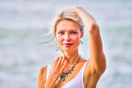 Beautiful young blonde blue eyes woman posing outdoor at the rocky sea shore. Trendy fashion female model dressed in white topの写真素材