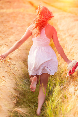 Happy young blonde Scottish girl in white dress with straw hat running away through the golden wheat fieldの写真素材