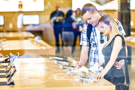 Young couple in consumer electronics retail store browsing at displayed latest tablets, choosing new...