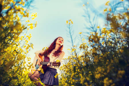 Young ginger hair girl in 70s style with acoustic guitar in rapeseed field.の写真素材