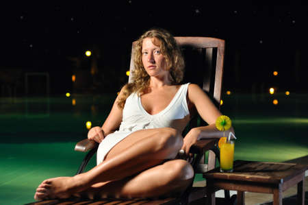 sensual woman is sitting on chair at poolside at night and looking at cameraの写真素材