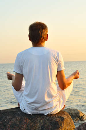 young man is sitting on a stone and looking at sea sunrise. view from behindの写真素材